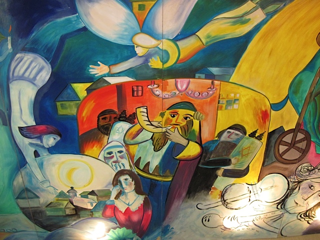 Colorful mural in the style of Marc Chagall depicting a shofar blower, a figure reading Torah, and community members gathered beneath a hovering angel.